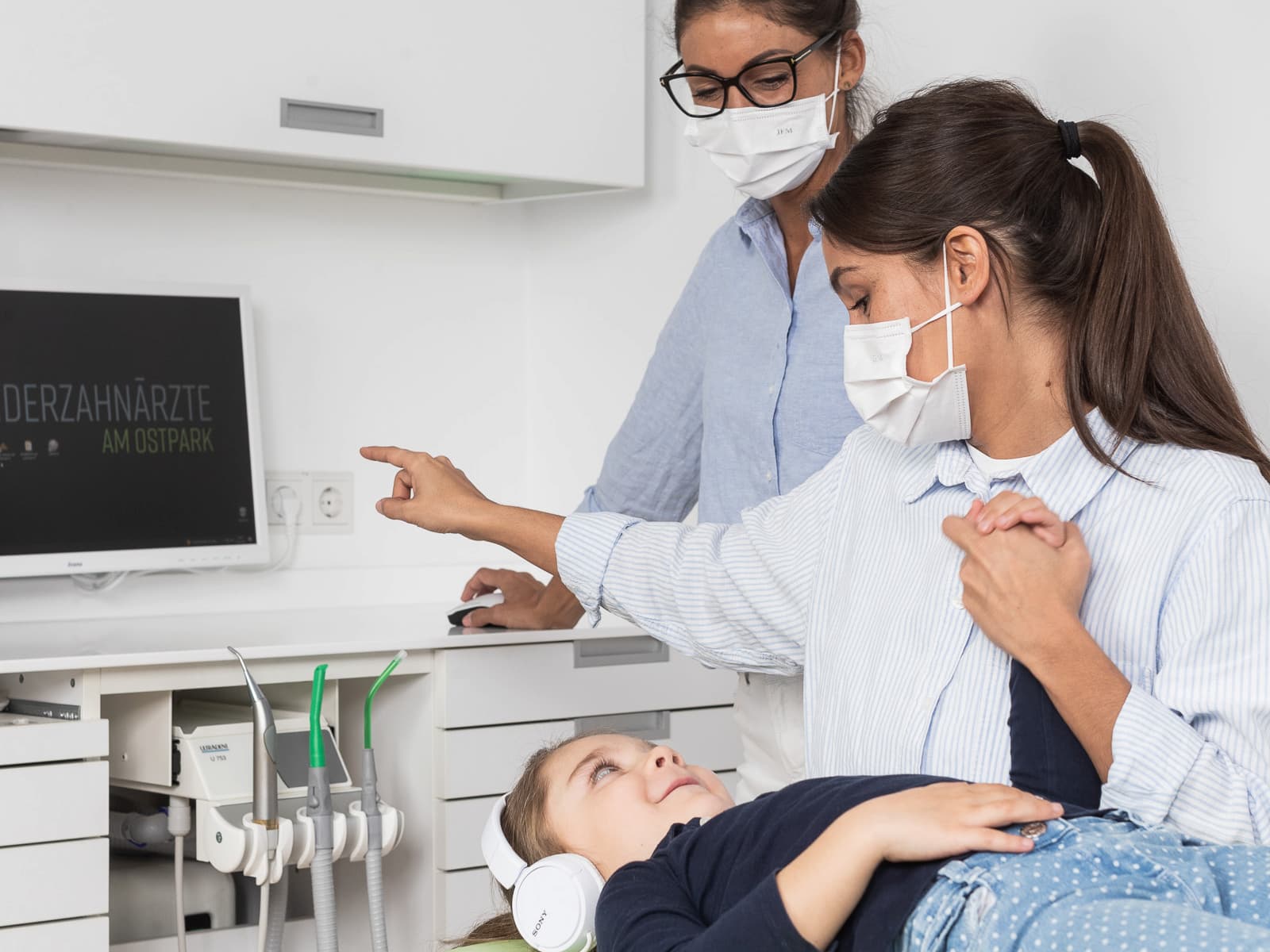 In einer Kinderzahnarztpraxis liegt ein Kind mit Kopfhörern entspannt auf dem Behandlungsstuhl, während eine Zahnärztin seine Hand hält. Eine zweite Zahnärztin zeigt auf den Bildschirm im Hintergrund, auf dem „Kinderzahnärzte am Ostpark“ steht.