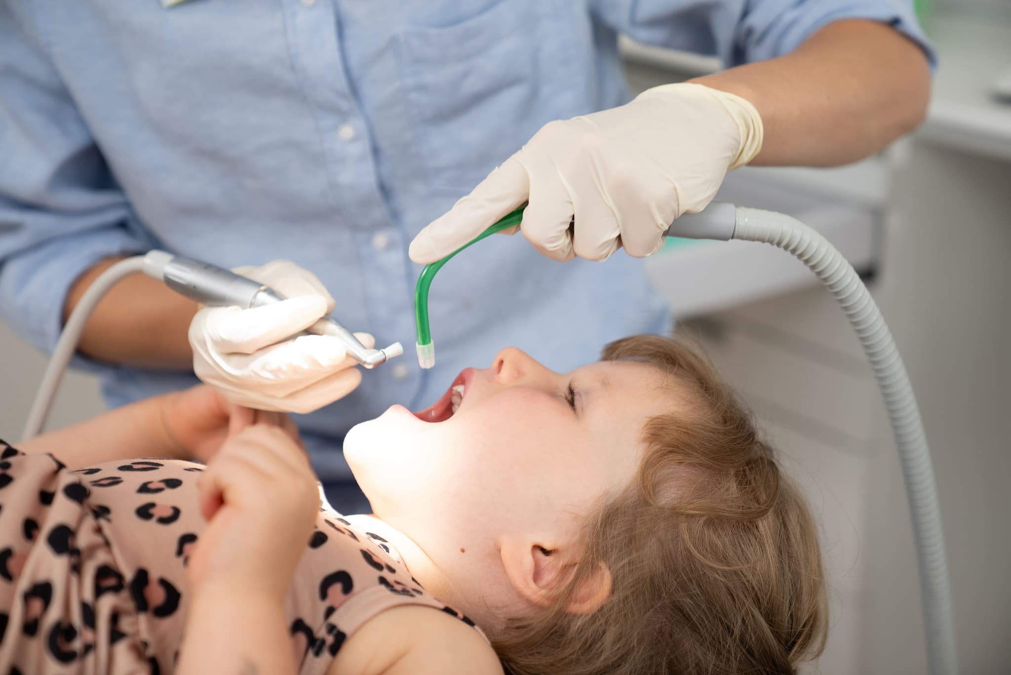 Ein Kind liegt mit offenem Mund auf dem Behandlungsstuhl, während eine Zahnärztin mit Handschuhen zahnärztliche Instrumente im Mund des Kindes anwendet.