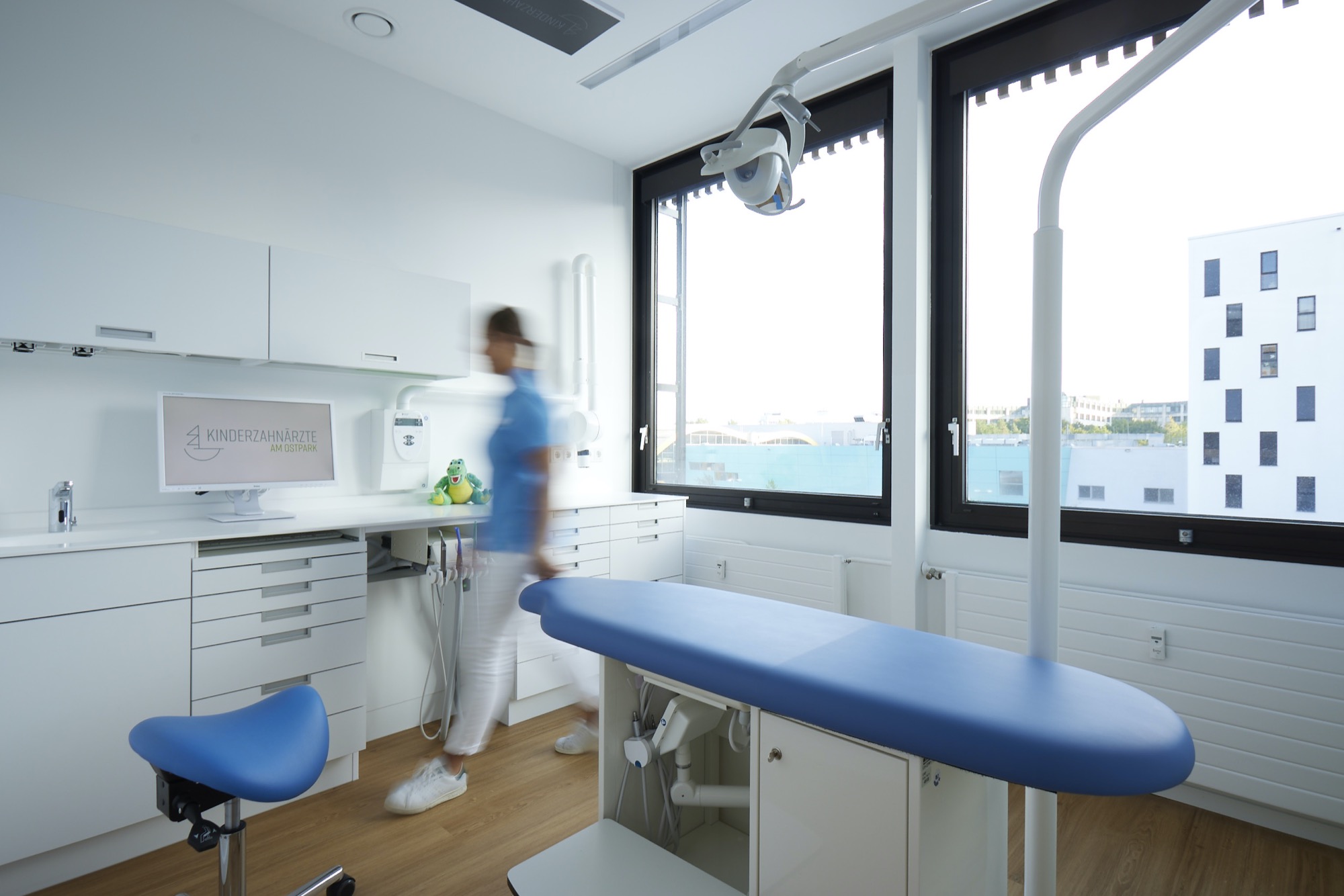 Das Bild zeigt ein helles, modernes Behandlungszimmer mit großem Fenster und kindgerechter Ausstattung. Eine Mitarbeiterin bewegt sich durch den Raum, was eine freundliche und lebendige Atmosphäre vermittelt.