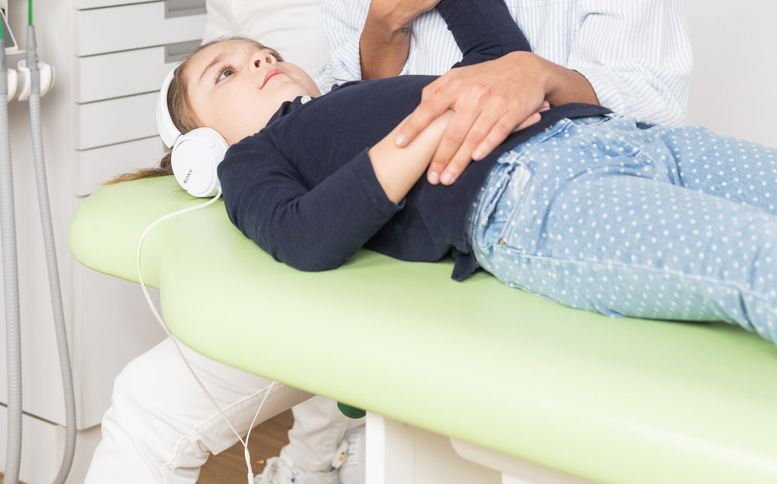 Ein Kind liegt mit Kopfhörern auf einem grünen Behandlungsstuhl, während eine erwachsene Person beruhigend eine Hand auf seinen Oberkörper legt.