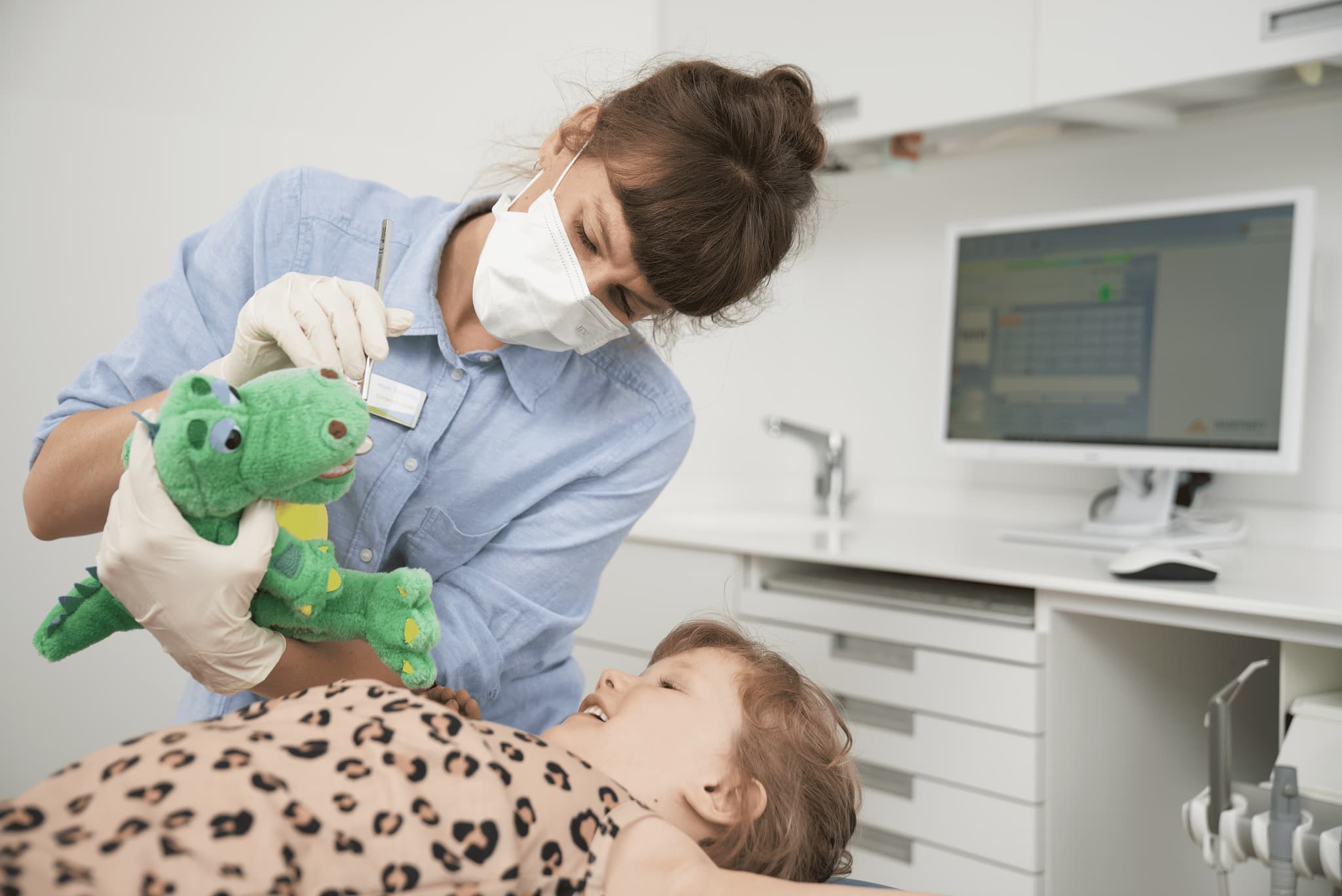 Eine Zahnärztin mit Maske und Handschuhen zeigt einem Kind spielerisch an einem grünen Stofftierkrokodil, wie die Behandlung abläuft. Das Kind liegt auf dem Behandlungsstuhl und schaut interessiert zu.