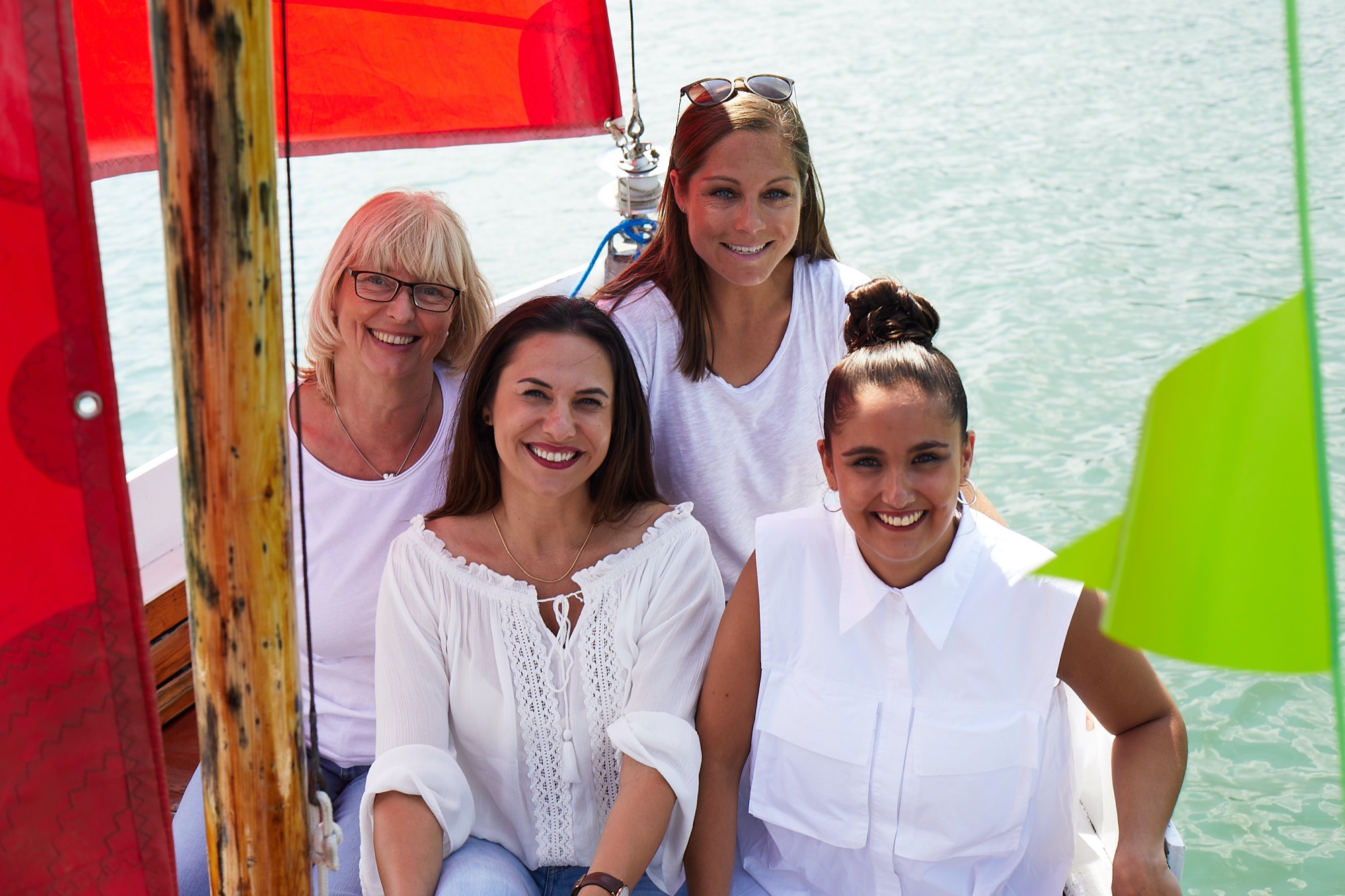 Vier Frauen sitzen lachend auf einem Boot mit roten Segeln, während im Hintergrund das Wasser zu sehen ist.